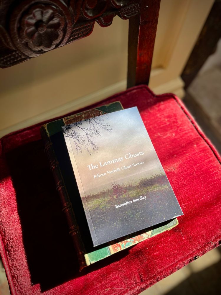 A small pile of books sits on a mid seventeenth century chair with a red velvet seat cushion. The top book is called The Lammas Ghosts: Fifteen Norfolk Ghost Stories by Barendina Smedley. Light falls on it from a window, unseen, to the right of the photo. The long shadows give the image an autumnal feeling. 
