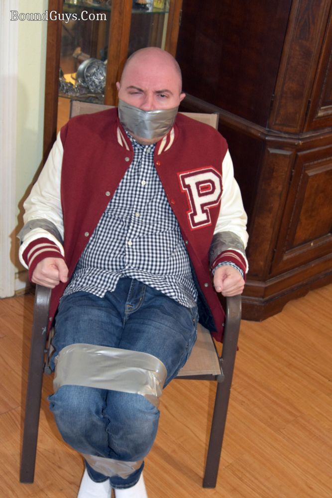 Model Casey duct taped arms, legs, and mouth. He's tied to a chair and wearing a red and white letterman jacket with a P on it.