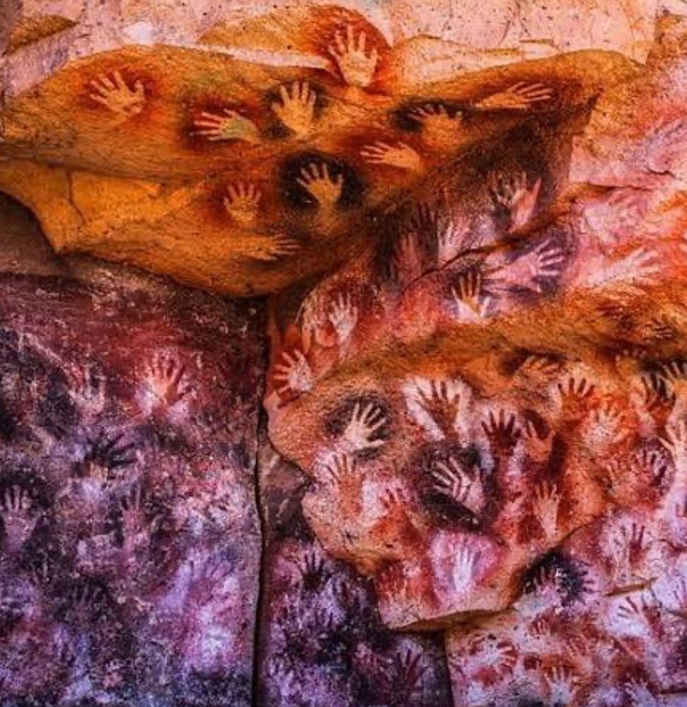 A imagem mostra um detalhe de uma parede rochosa que exibe centenas de estênceis de mãos humanas. As impressões das mãos são de várias cores, incluindo tons de branco, preto, vermelho, marrom e roxo, e estão sobrepostas umas às outras, criando uma textura densa e visualmente complexa. A rocha em si apresenta cores quentes e terrosas, com áreas de laranja, vermelho e marrom. A iluminação parece ser natural, realçando as texturas e as cores da superfície.