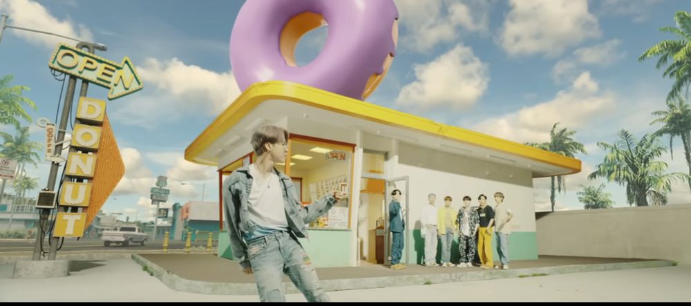 Screenshot from the MV for BTS’s song Dynamite. Jimin is dancing in the parking lot of a donut shop with a giant pink iced donut on the roof. The other 6 members are standing in the background near the door to the shop watching him. Jungkook is holding a donut. 