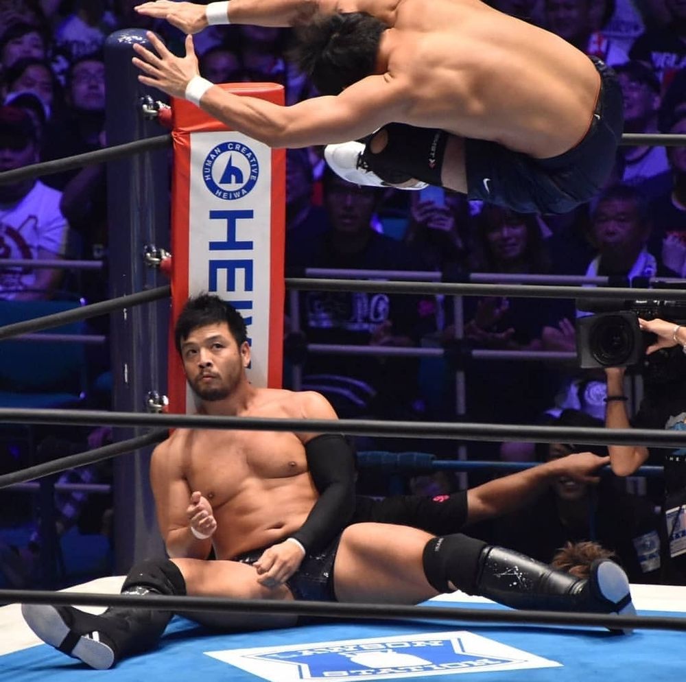 KENTA seated against a turnbuckle pad while Katsuyori Shibata, in midair, is about to dropkick him in the head.