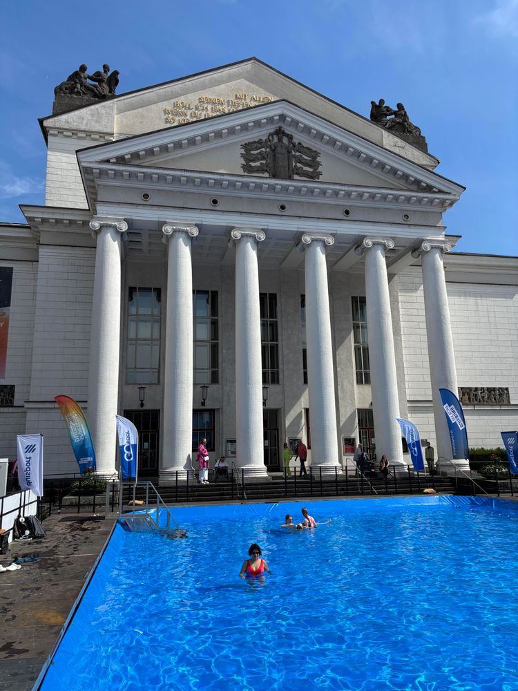 Ein großes Schwimmbecken ist vor dem Duisburger Stadttheater aufbaut. Man sieht die Säulen des Gebäudes im Hintergrund und das blaue Becken mit Schwimmer:innen