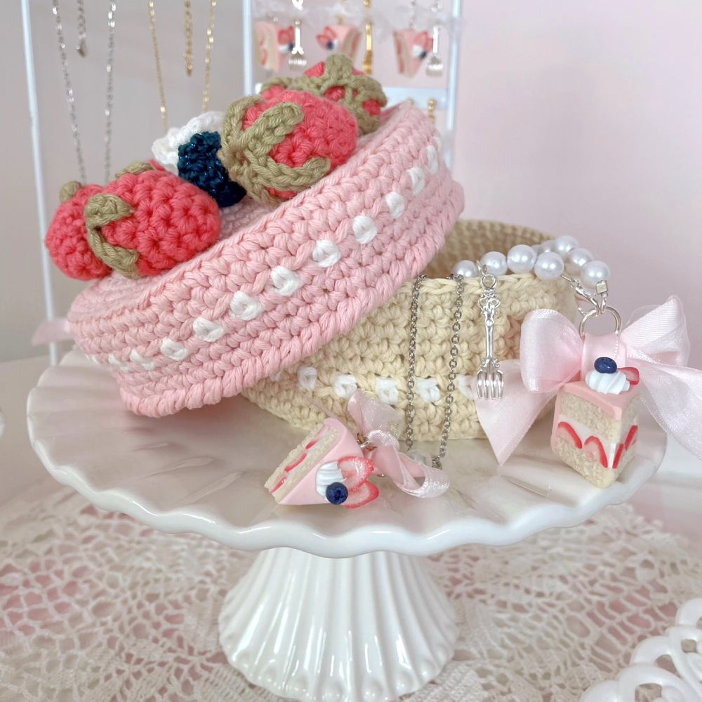 Photo of a crocheted jewelry box in the shape of a pink cake with berries on top. A necklace and charm are positioned in front of the jewelry box, both in the shape of slices of pink cake.