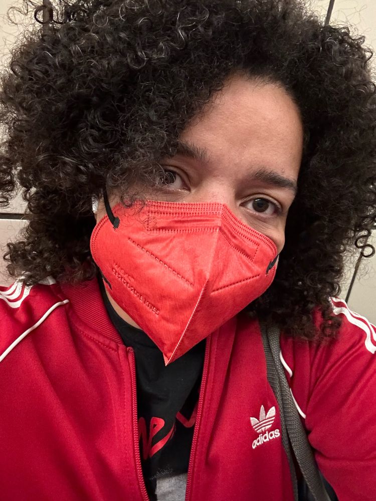 A selfie of a person of colour wearing a red mask and red ADIDAS jacket looking into the camera. They also have on AirPods and have very curly dark hair.