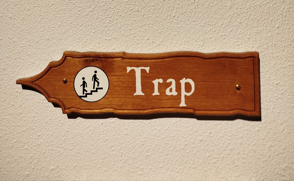Ein Holzschild an einer weißen Wand, zeigt nach links, mit einem Treppensymbol und dem Wort Trap in weiß geschrieben 
