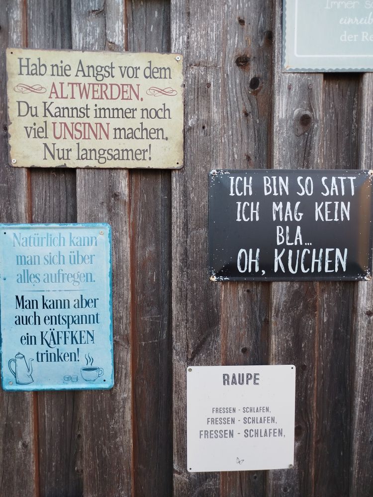 Eine dunkle Holzwand mit vier Schildern.
"Hab nie Angst vor dem Altwerden, du kannst immer noch viel Unsinn machen. Nur langsamer!"
"Ich bin so satt
Ich mag kein Bla... Oh, Kuchen"
"Natürlich kann man sich über alles aufregen. Man kann aber auch entspannt ein Käffken trinken!"
"Raupe
Fressen - Schlafen 
Fressen - Schlafen 
Fressen - Schlafen"