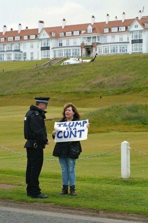 Janey being spoken to by a police officer while holding her Trump Is A Cunt sign in front of Trump's golf course