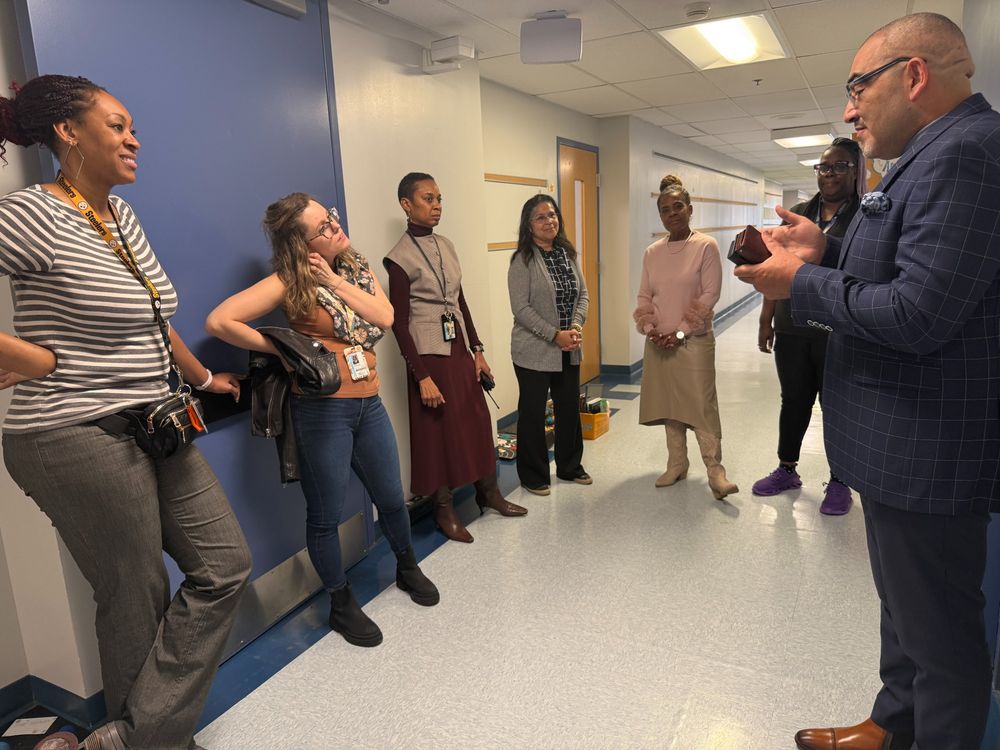 Dr. Francisco Durán stands in a school hallway, speaking to a group of six staff members. The staff, dressed in a mix of casual and professional attire, listen attentively and smile. One staff member leans against the wall while another stands with hands clasped.