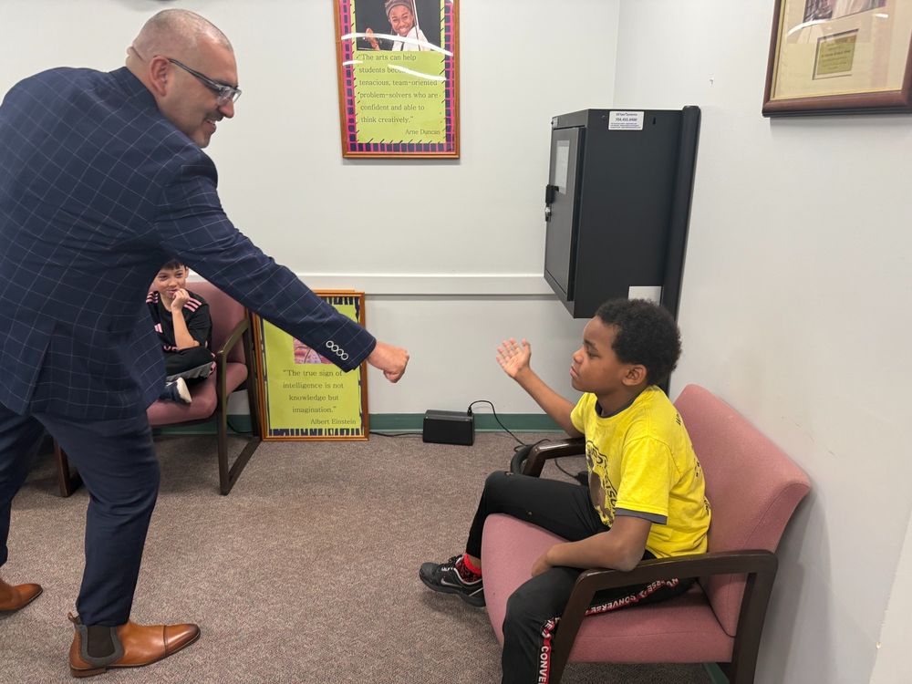 Dr. Francisco Durán leans in with a friendly fist bump toward a student wearing a yellow shirt, who sits in a chair with an outstretched arm. Another student in a black and pink jersey sits nearby, watching with a smile. A motivational poster featuring an Albert Einstein quote is visible on the wall behind them.