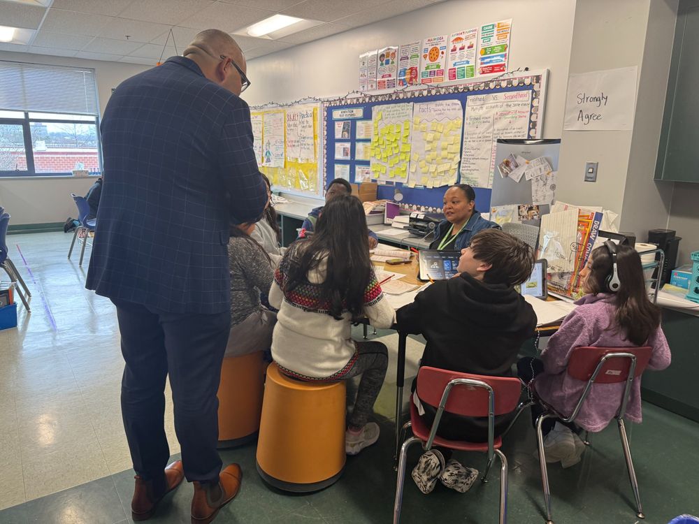 Superintendent Dr. Francisco Durán stands in a classroom, wearing a dark blue checkered suit, engaging with a small group of students working at a table. A teacher sits with the students, smiling as they interact. The classroom walls are decorated with educational posters and sticky notes.