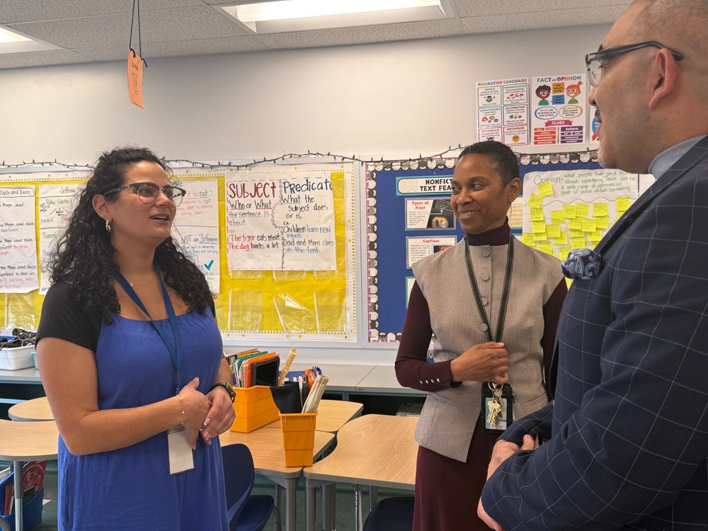 Dr. Francisco Durán speaks with a teacher in a classroom alongside the principal. One teacher wears a blue dress and glasses, smiling as she talks, while the other wears a gray vest over a burgundy top, listening attentively. The background features a classroom bulletin board covered in sticky notes and posters related to reading and writing skills.