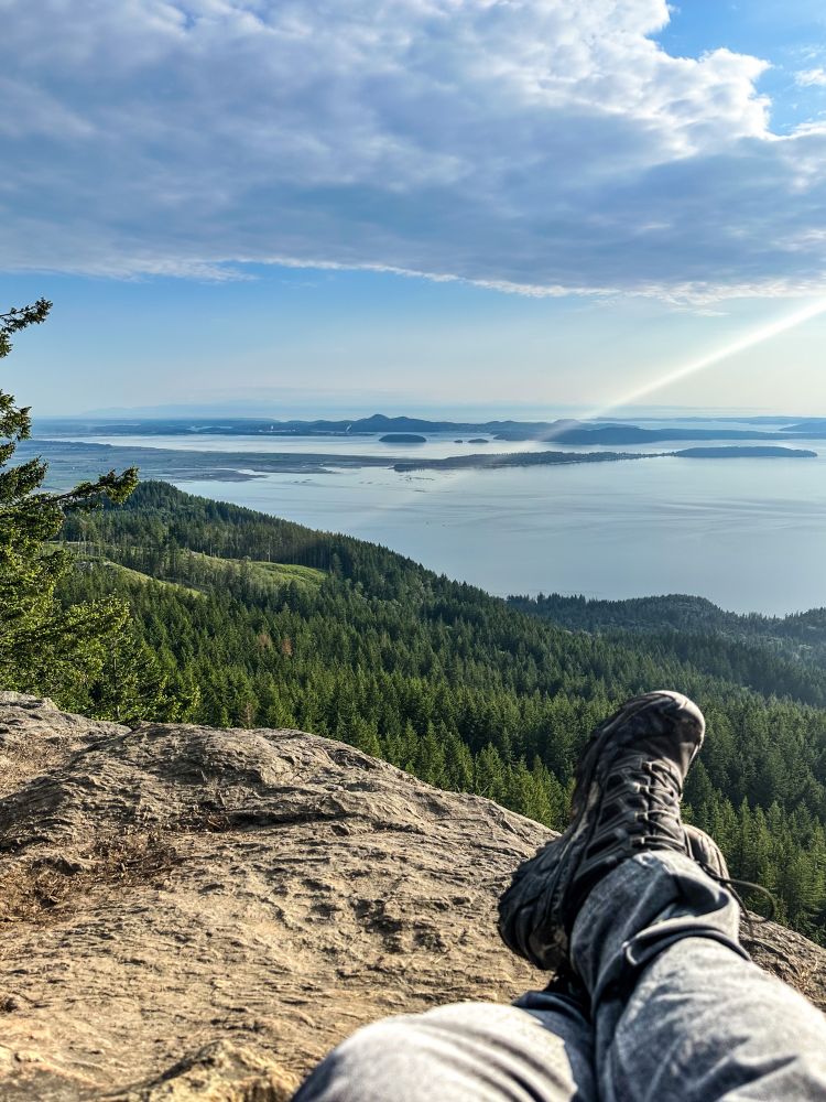 On a mountaintop overlooking a lake, tall redwood trees near the lake and on the mountains, my legs and hiking boots in the front of the camera 