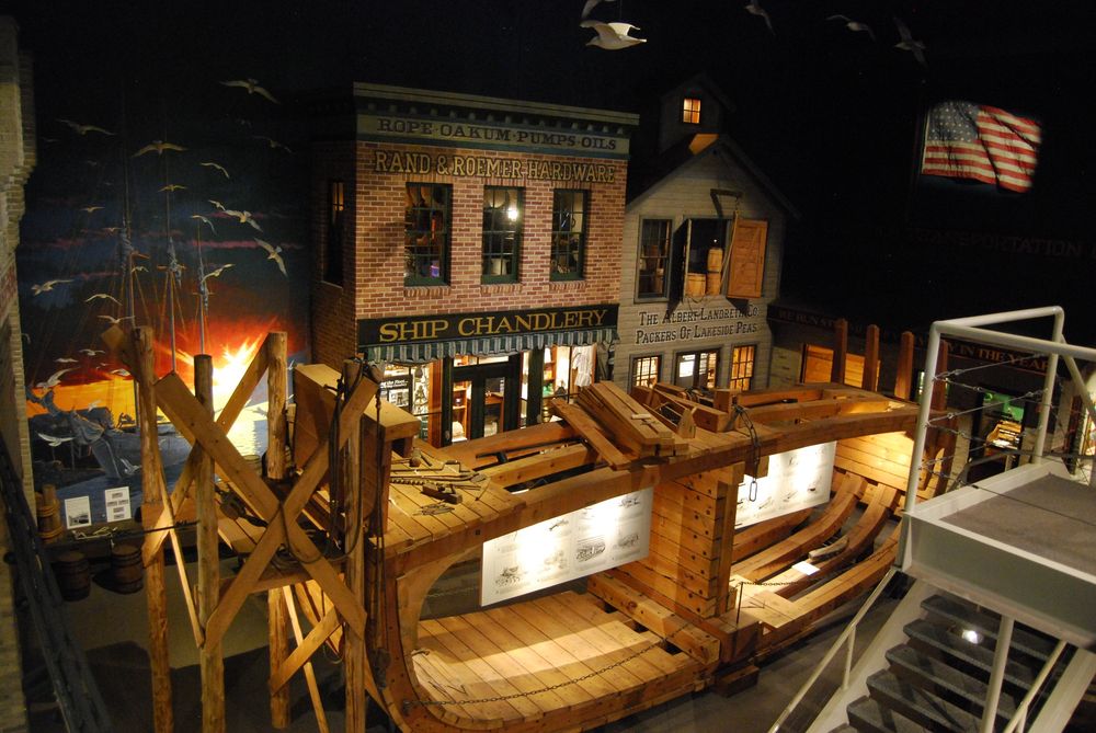 An elevated look into a downstairs section of the maritime museum, containing false building facades that look like old shipbuilders, and a large wooden section of a ship hull.
