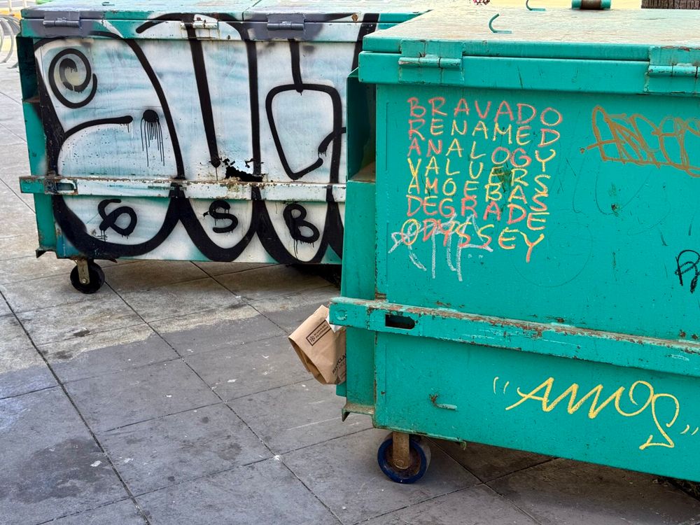 Two dumpsters on a city sidewalk with graffiti on them. One of them has 7 words written in pink/yellow marker. The words form a square and can be read horizontally or vertically.