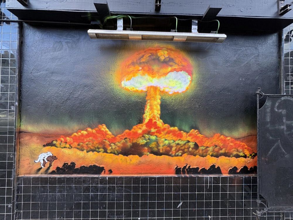 A mural depicting a mushroom cloud from a recent explosion. It looks like a nuclear bomb maybe, with dark clouds but orange and yellow light emanating from the explosion like a tiny sun. In the left-hand corner, two dogs are fucking. They are unbothered by the nearby explosion and danger.