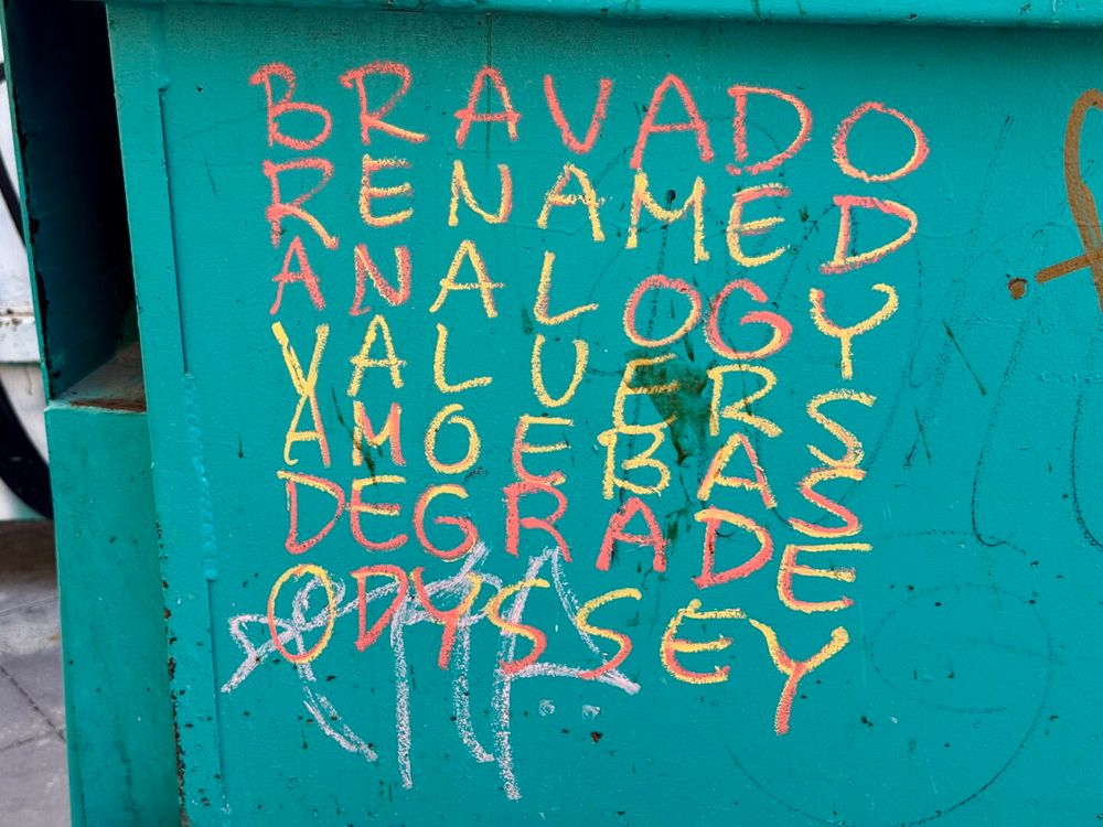 An acrostic poem written in marker on a green dumpster. The "poem" consists of 7 words written on seperate lines. Each word has the same number of letters and are written in a way that allows the reader to find the same word written vertically or horizontally. So the first word is Bravado, but if you right the first row of letters from top to bottom, you'd also see "Bravado". The words are: Bravado, Renamed, Analogy, Valuers, Amoebas, Degrade, and Odyssey.