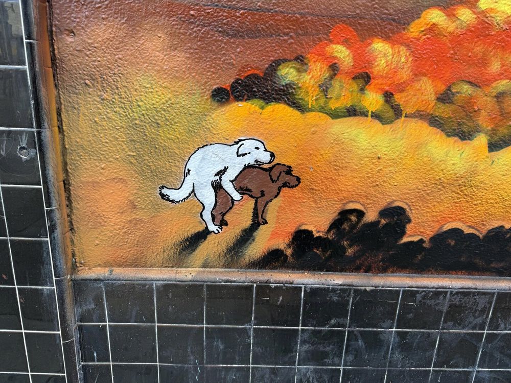 A closer view of two dogs humping in the lower left-hand corner of the previous mural. One dog is white, the other black. Neither seems bothered by the nearby nuclear explosion, even as the light from it is bright enough to cast a heavy shadow on the ground of the two dogs fucking.