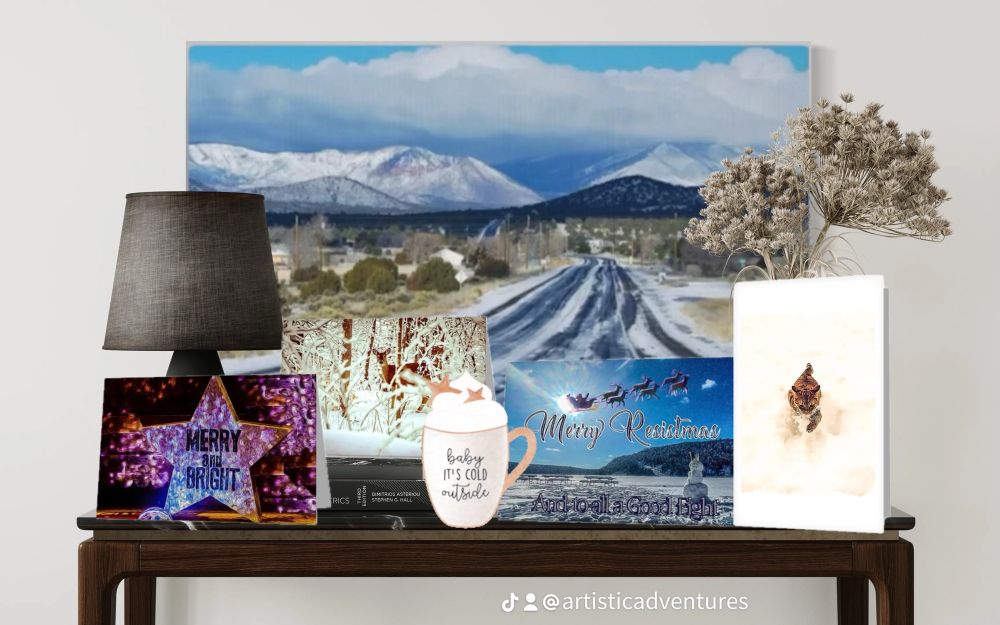 A framed landscape print hanging on a wall: a snowy mountain road leading toward a dramatic blue–white ridge under a wide winter sky. A small table beneath it holds candles, a plant, and holiday décor including gift cards from Artistic Adventurers holiday collection.