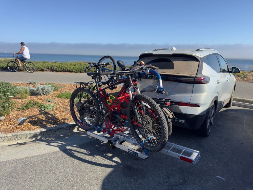 A silver Chevy Bolt compact EV with a trailer mount bike hitch carrying three bikes. In the background, Monterey Bay on a moderately sunny day.