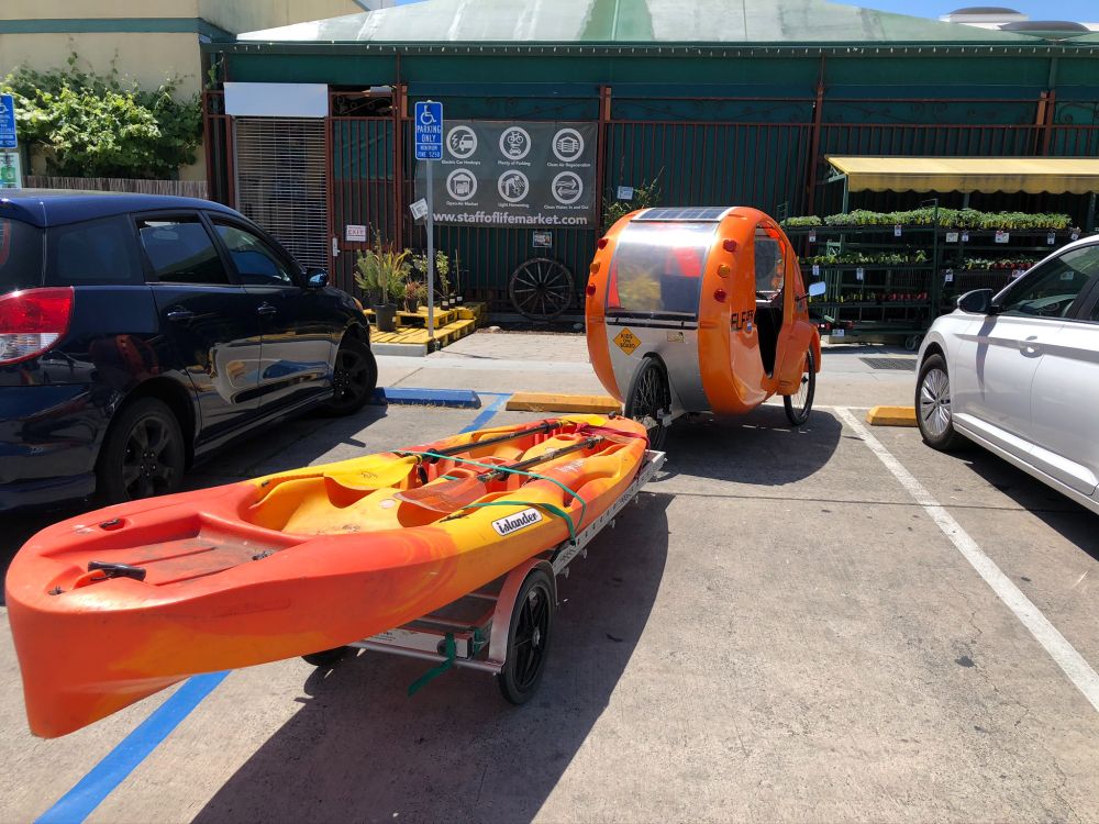 An orange velomobile (e-trike with a partial enclosure for sun+rain protection) with an aluminum trailer, on which is an orange double kayak. It's parked in a parking spot at a grocery store.