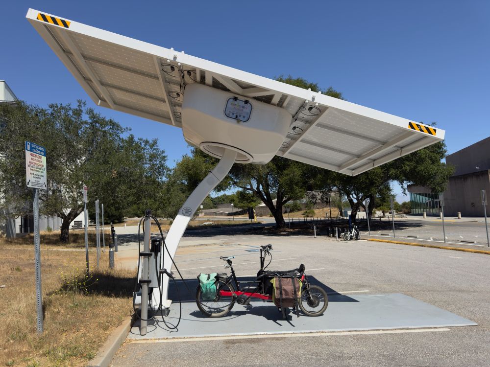 A side view of a bakfiets e-bike plugged in at a solar EV charging station in a parking lot.