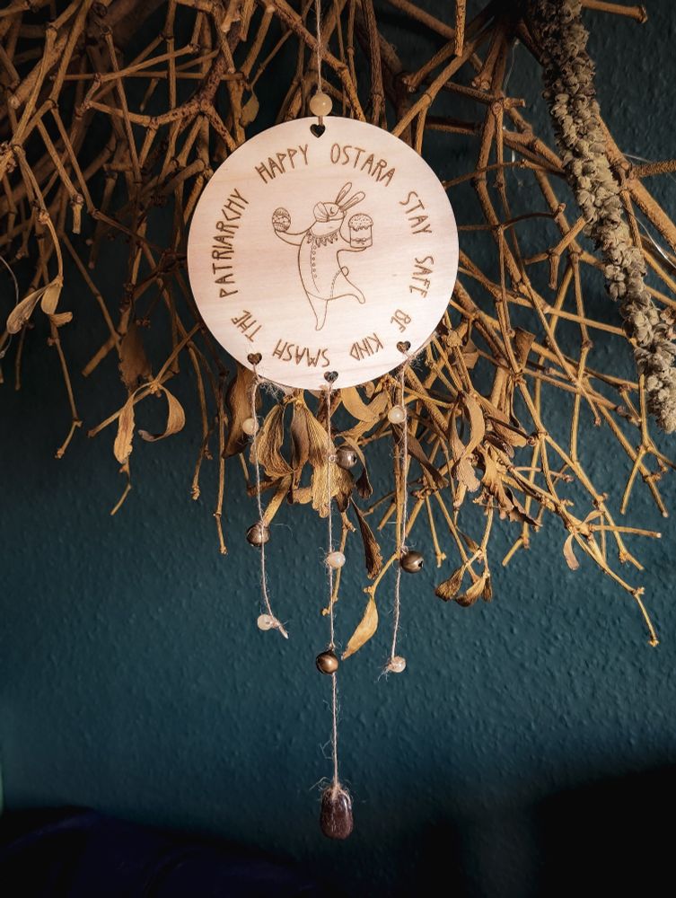 An einer getrockneten hängenden Mistel baumelt eine Holzplatte an einer Schnur. Unten an der Holzplatte hängen drei Juteschnüre die abwechselnd mit gelblichen, braunen Steinen und Glöckchen bestückt sind. In der Holzplatte ist ein Motiv und Schrift eingebrannt. In der Mitte ist ein gekleideter Hase zu sehen, der in einer Hand ein Ei und in der anderen ein Törtchen trägt. Außerdem hat er eine medizinische Maske auf.
Um ihn herum steht kreisrund der Text: Happy Ostara stay safe be kind smash the patriarchy.