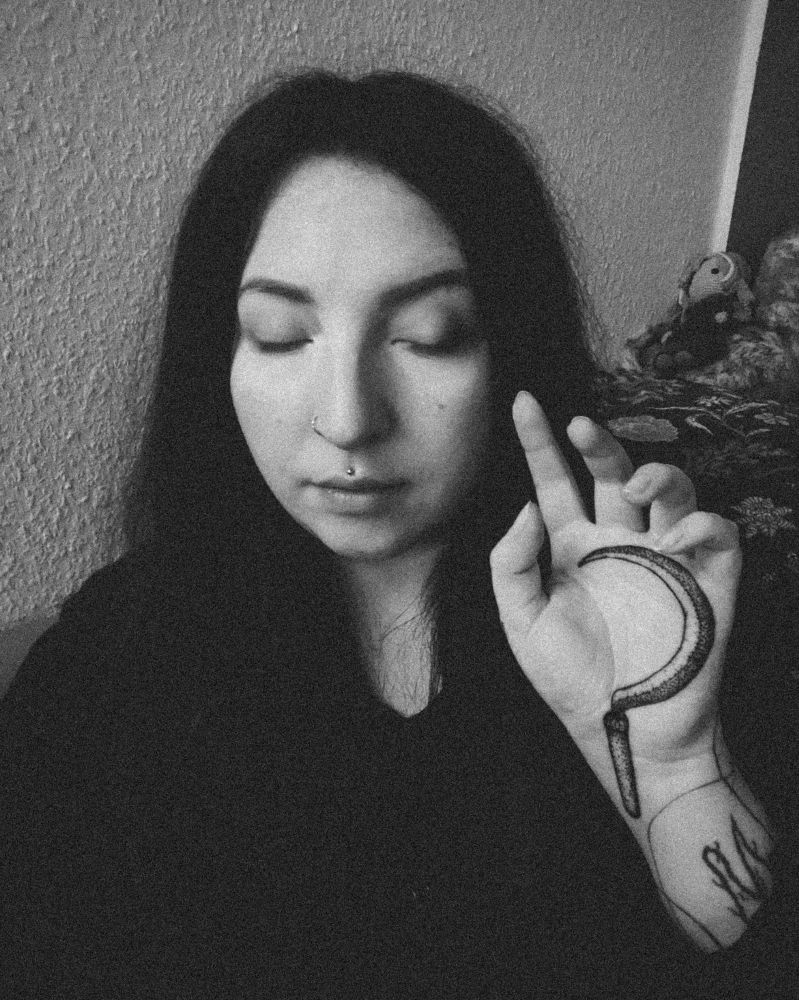 Ein schwarz-weiß Portrait von mir. Meine Augen sind geschlossen und ich habe einen friedlichen Gesichtsausdruck. Ich hebe eine meiner Hände ungefähr auf Kopfhöhe, sodass meine Handinnenfläche sichtbar ist. Auf meiner Handinnenfläche ist ein Tattoo, das eine Sichel zeigt.