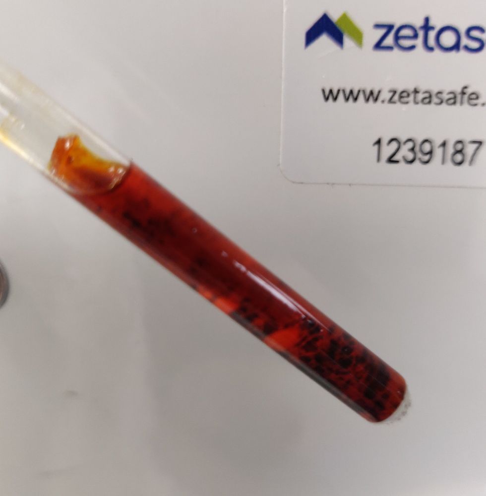 A photo of an NMR tube, with a dark brown solid, with some much larger patches of orange present compared to the previous photo