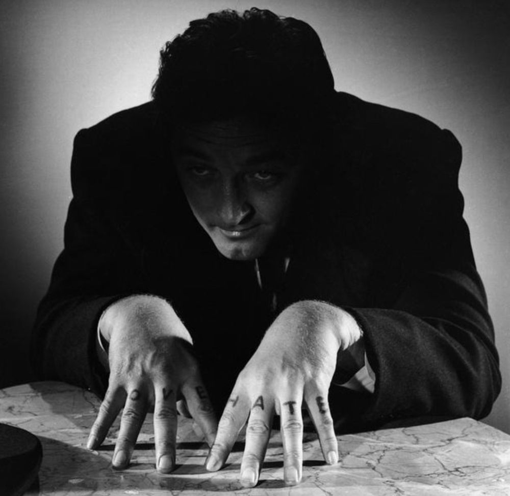Robert Mitchum as Harry Powell in Night of the Hunter with steepled hands, fingertips touching a marbled surface. He sits partially in darkness
