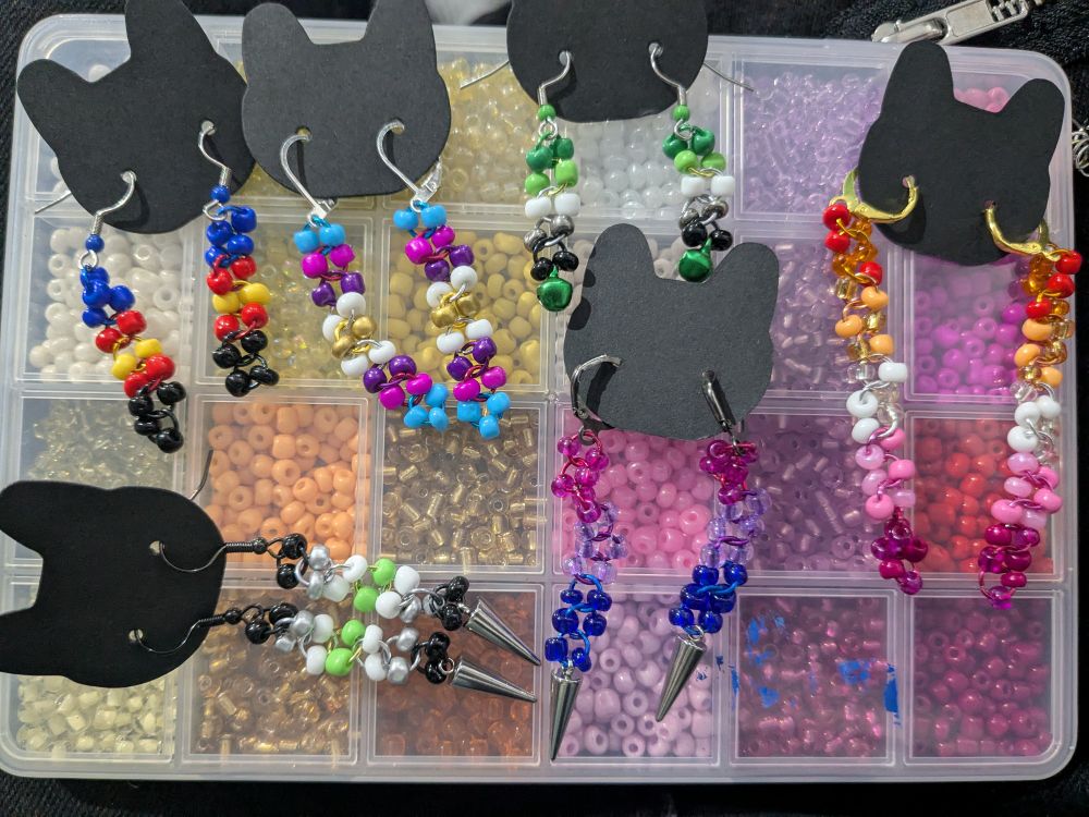 a variety of pride earrings, patterns include lesbian, bisexual, agender, aromatic, and two polyamory pride flags made with jump-rings and seed beads