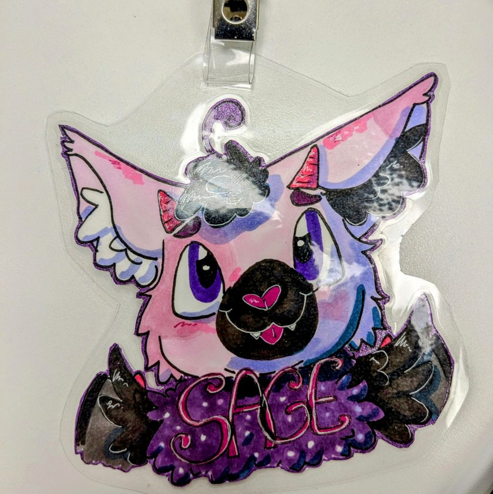a laminated furry badge of a sona named Sage. she is a pink cat bat with horns
