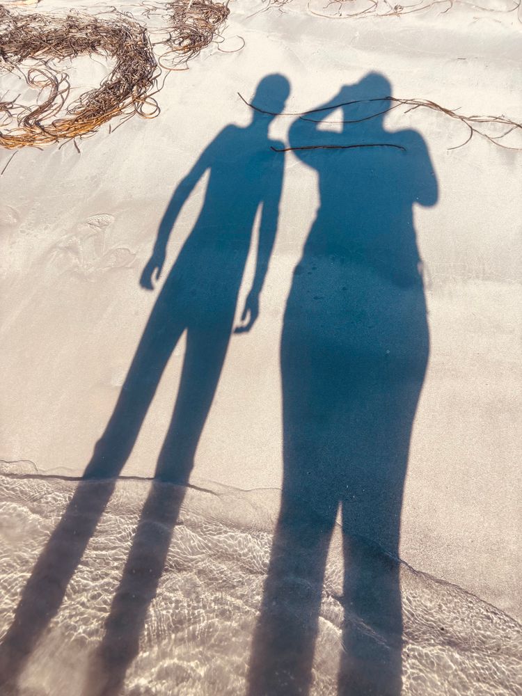 A photograph of our two shadows as we come out of the sea from a swim, with sunny ripples on the shore and the beach #SonAndMother 