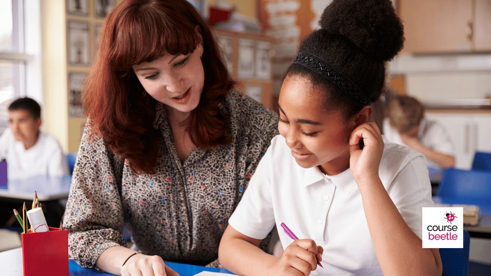 A teacher sits next to a girl in her class at a table. The teacher supports the child who has hearing loss.