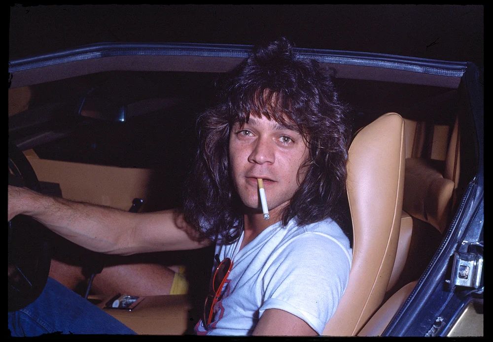 Eddie Van Halen with long hair (probably in his mid twenties) and in the drivers seat of a t-top Camaro. His hand is on the steering wheel and he has a cigarette dangling from his mouth. The picture is epic rick and roll. 