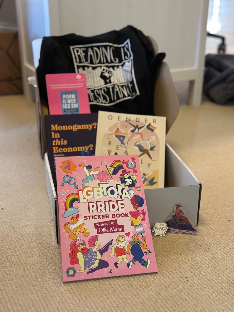 A collection of books and pins are propped up against a cardboard box that they were mailed in. A black t-shirt with “reading is resistance” and an illustration of a fist in white, “LGBTQIA+ Pride Sticker book”, the book “Monogamy? In this economy?”, a card that says “Porn is not sex ed” and a partially obscured cover of another book 