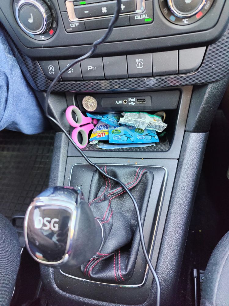 A photograph of a car's interior where the stick is, showing a little tray stuffed with glass wipes, mints and scissors. Above the tray is a St. Christopher's coin which came with the car. And there's an audio cable-in which reads: AUX/iPod and a USB port next to it.