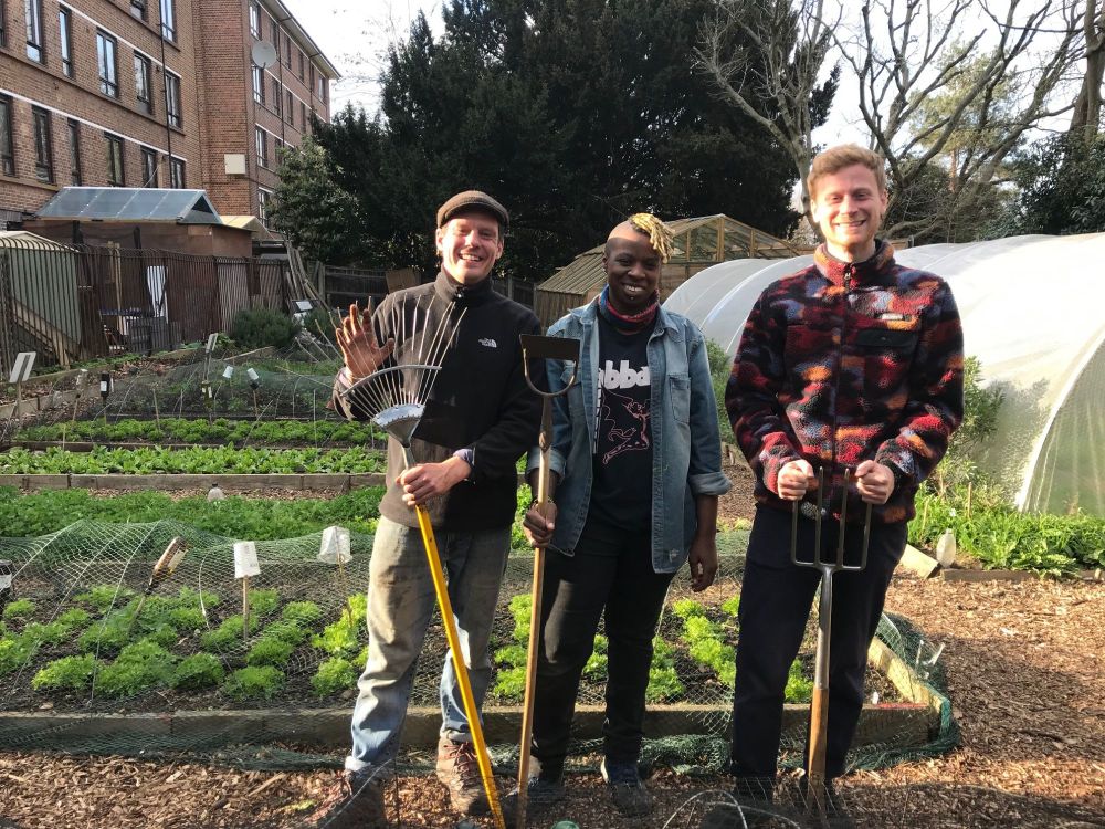 Three smilling young people holding gardening tools in front of food growing beds