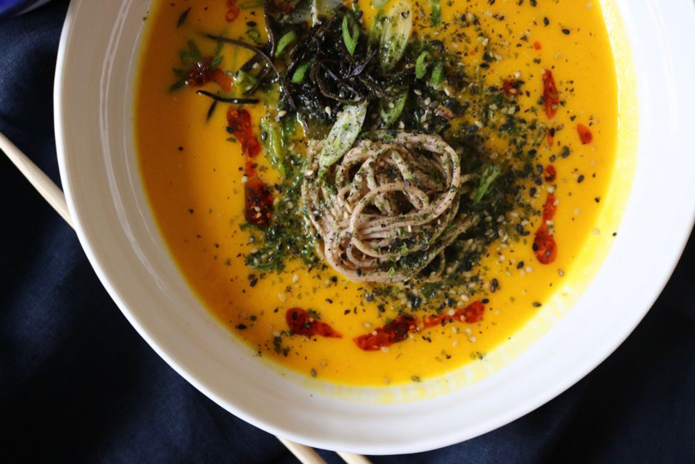 white bowl filled with yellow coloured soup, noodles and green veg with drizzled chilli sauce by Kali Jago
