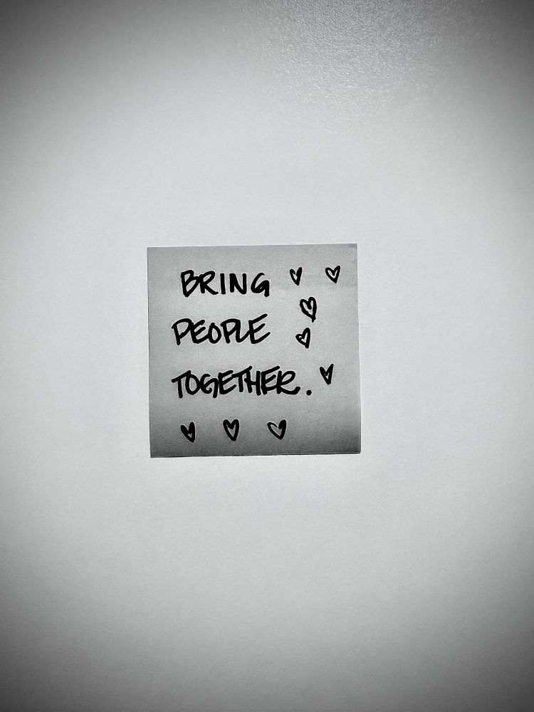 Black and white photo of a post it that reads: “Bring People Together” with hearts drawn around it. 
