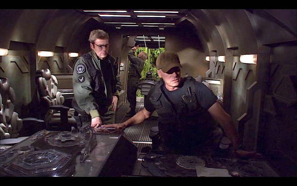 Daniel and Jack are inside a spaceship. Daniel, wearing green BDUs and glasses, stand near Jack, who is wearing a brown cap, black T-shirt and vest. He's seated on the pilot's chair. 