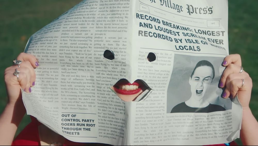 ur mum, by wet leg. music video. screenshot of the main singer behind a newpaper cutout for her mouth. headline says record breaking: longest and loudest scream ever recorded by isle of white locals, referring to the scream she does in this song which is quite long and long indeed