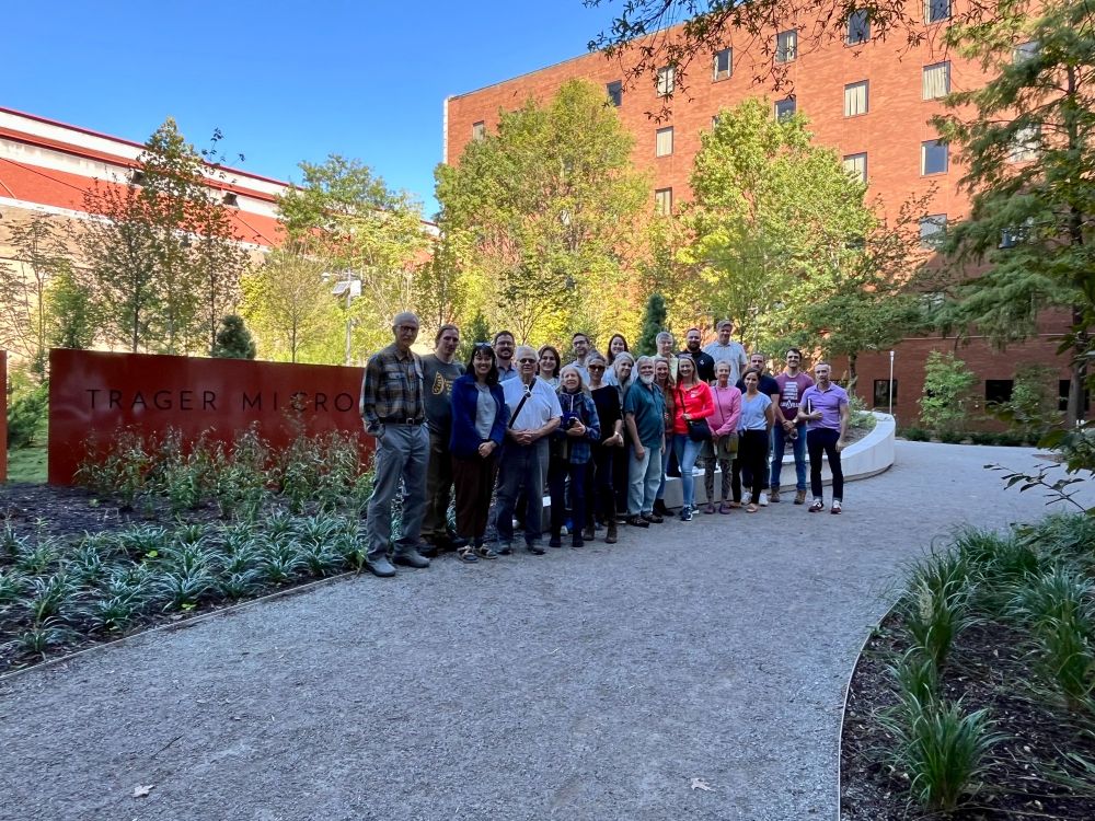 Group photo from the Trager MicroForest tour on September 27, 2025. Group of about two dozen attendees stands on crushed stone path in front of Trager MicroForest sign with trees, city buildings and a bright blue sky in the background.