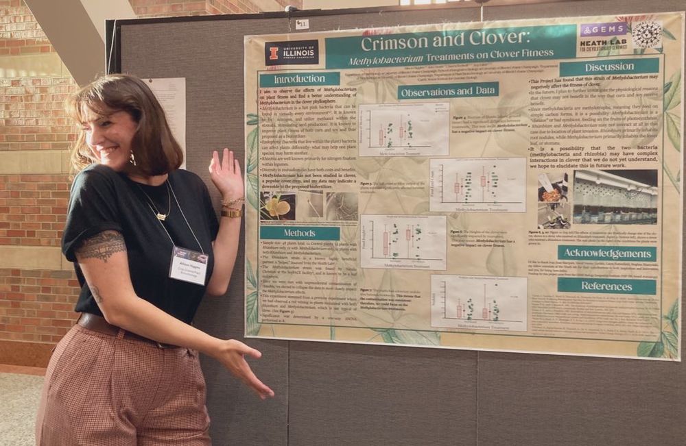 Allison (op) posing next to a research poster on the effects of methylobacterium on clover. She is wearing a black shirt and brown slacks, and gesturing toward the poster. The poster is green with text, graphs, and images of the research