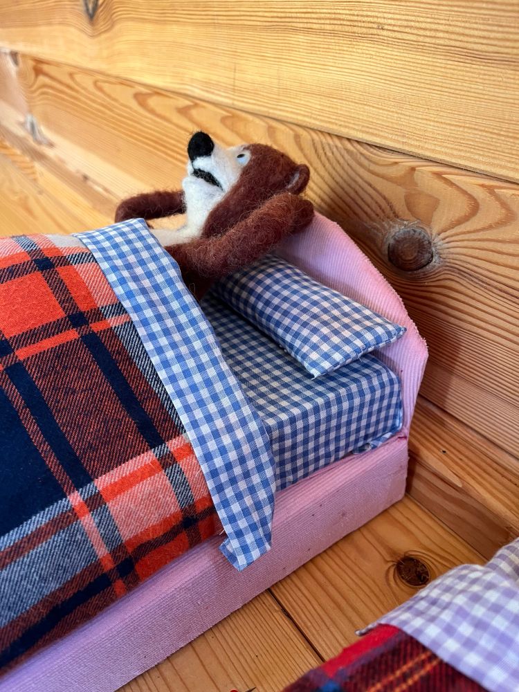 Close up of daddy bear stop motion animation puppet created in felt tucked up in bed. The bed is made of pink corduroy, the sheets are checked blue and white and the blanket is tartan style in blue and orange  