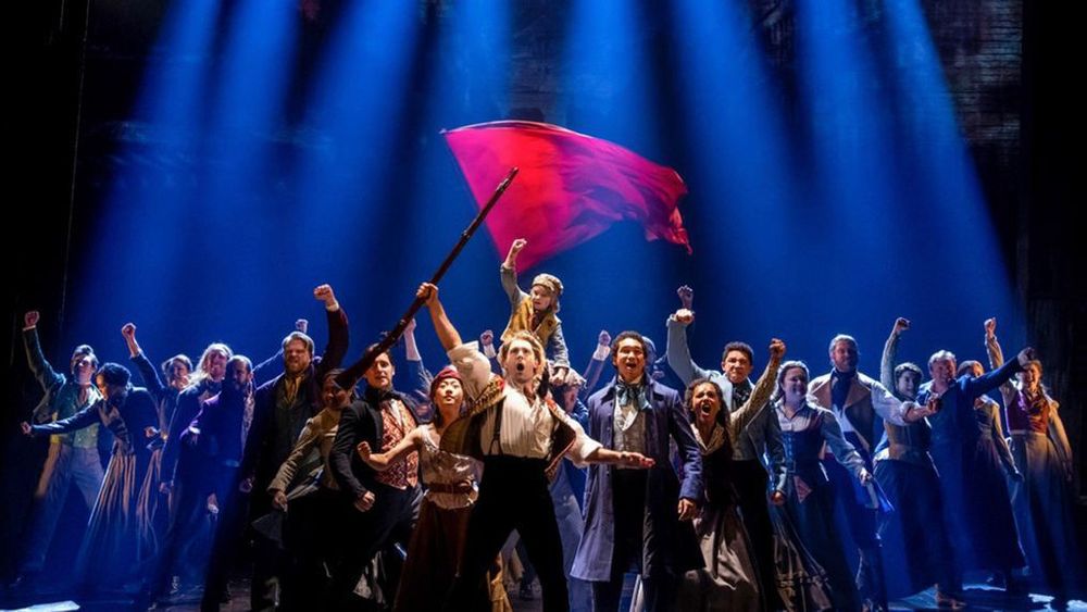 Photo shows the ending pose from One Day More from a stage production of Les Miserables. A crowd is assembled, fists raised. A red flag is in the center. 