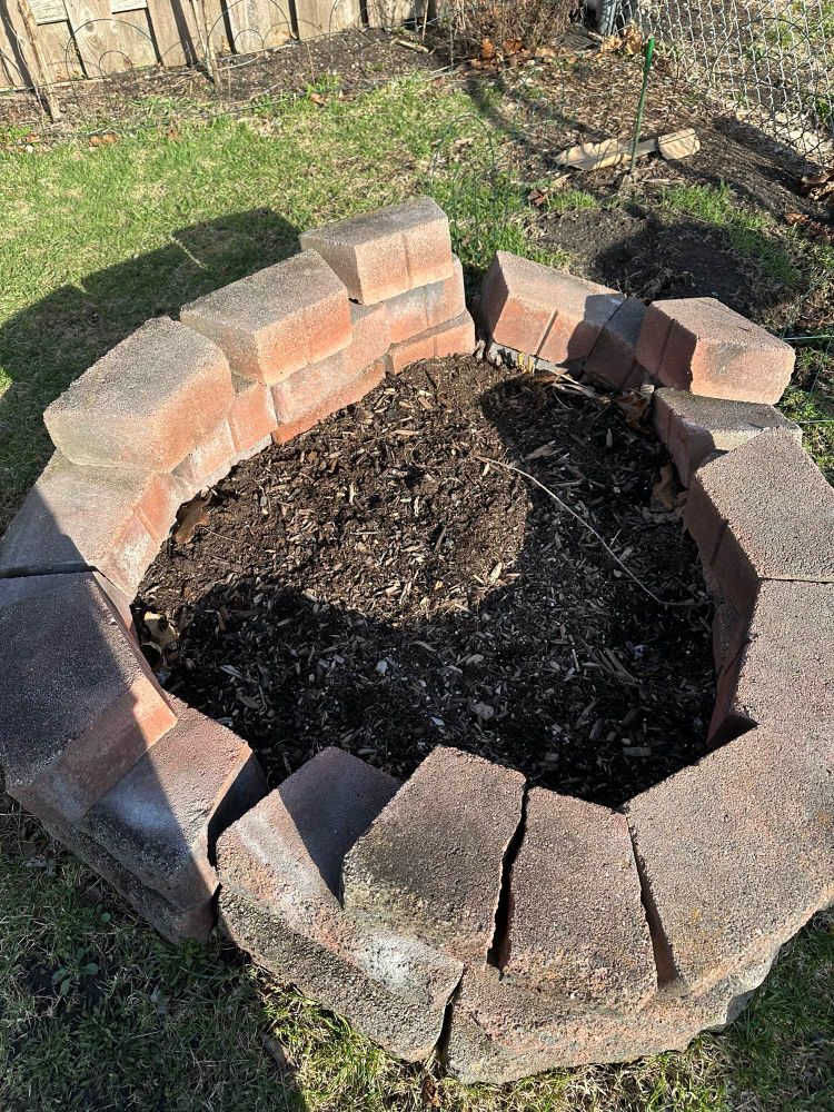 A circular homemade fire pit made of stacked trapezoid-shaped concrete pavers is filled with potting soil. 