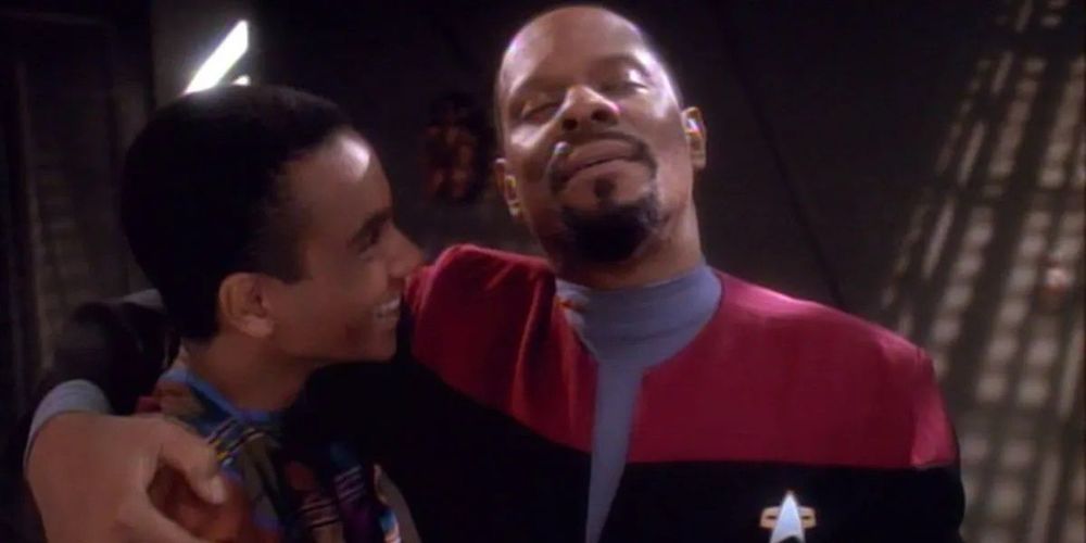 Photo is a still from Star Trek Deep Space Nine. Captain Sisko has his eyes closed and embraces his son, Jake with his right arm. Jake looks up at him and is smiling. 