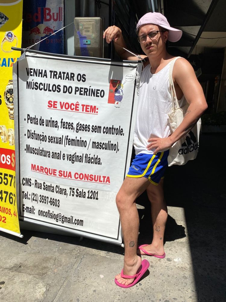 eu (homem branco cis, olhos e cabelo castanhos, boné lilás pro lado, oculos de grau com armaçao preta, regata branca, short azul, chinelo rosa e eco-bag) posando com a mão na cintura e um pé na pontinha ao lado de um cartaz que diz:

"VENHA TRATAR OS MÚSCULOS DO PERÍNEO
Se você tem:
- perda de urina, fezes, gases sem controle.
- disfunção sexual (feminino/masculino)
- musculatura anal e vaginal flácida

Marque sua consulta"
