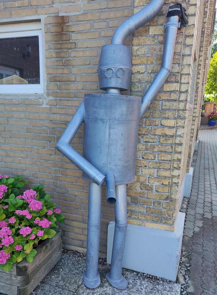 Regenfallrohr in Form eines Blechmannes, welcher lasziv an der Wand lehnt, bestückt mit einem Endstück, was wie ein Penis wirkt 