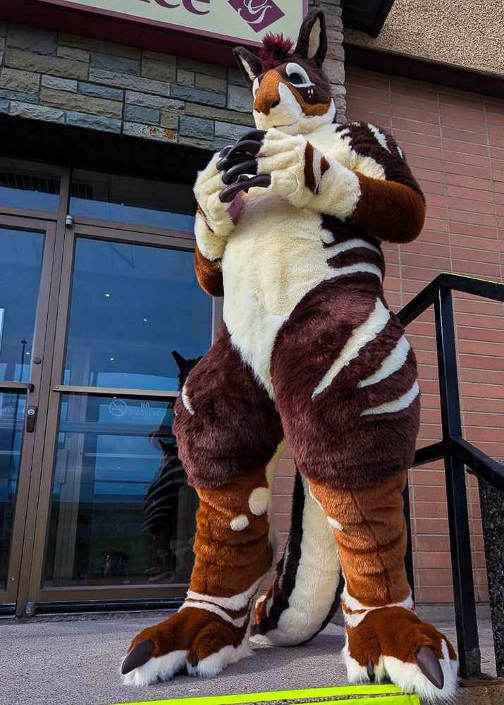 A kangaroo fursuit standing tall, with claws tapping against one another in front of his chest. He is standing in front of a hotel's exterior door, at the top of a set of stairs.